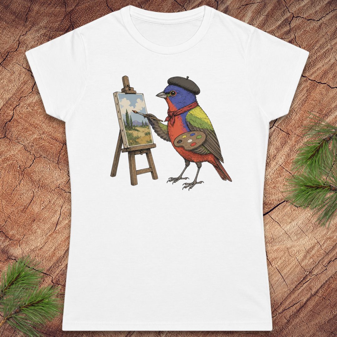 A white women's short-sleeve crewneck t-shirt featuring a colorful graphic of a bird painter on an easel, displayed flat on a rustic wood background with pine needles.