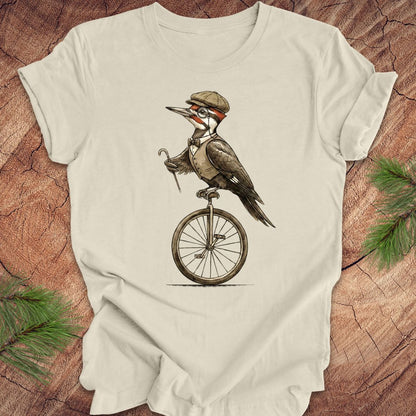 Beige t-shirt with a woodpecker on a unicycle graphic on a wooden surface.