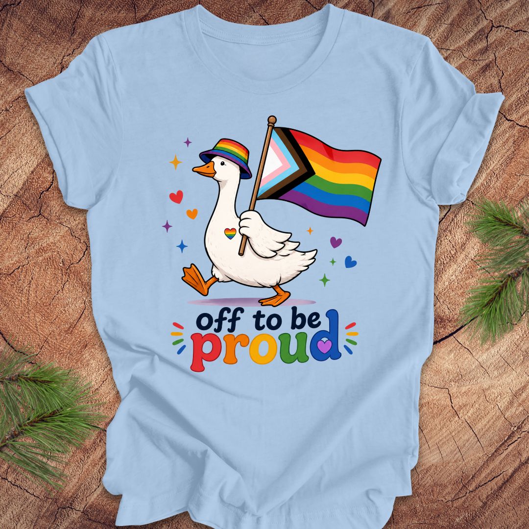 Light blue t-shirt with a goose holding a rainbow flag and text 'off to be proud' on a wooden surface.