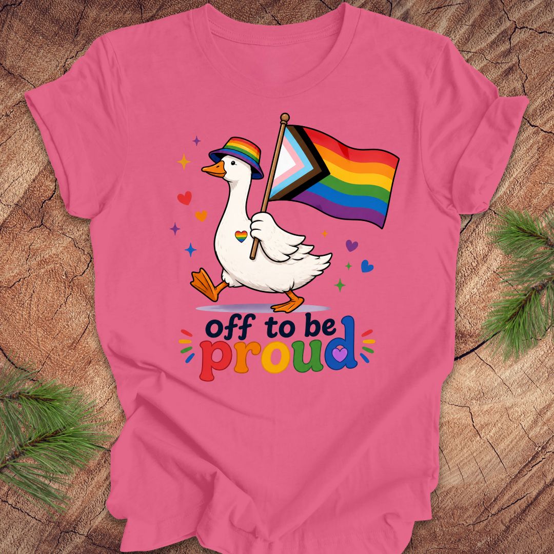 Pink t-shirt with a goose holding a rainbow flag and text 'Off to be proud' on a wooden surface.