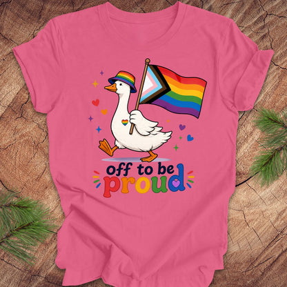 Pink t-shirt with a goose holding a rainbow flag and text 'Off to be proud' on a wooden surface.