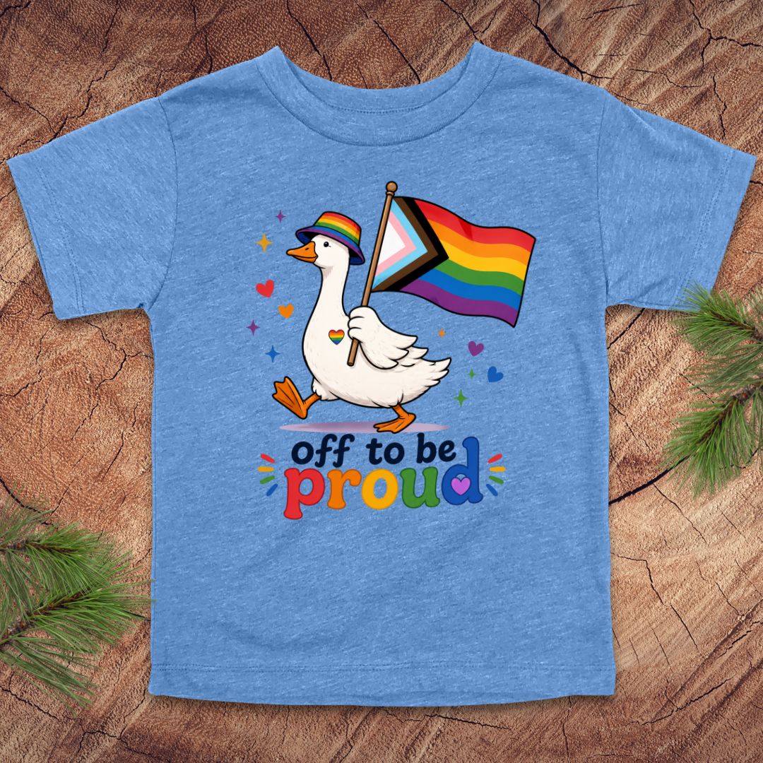 Blue t-shirt with a goose holding a rainbow flag and text 'off to be proud' on a wooden surface.