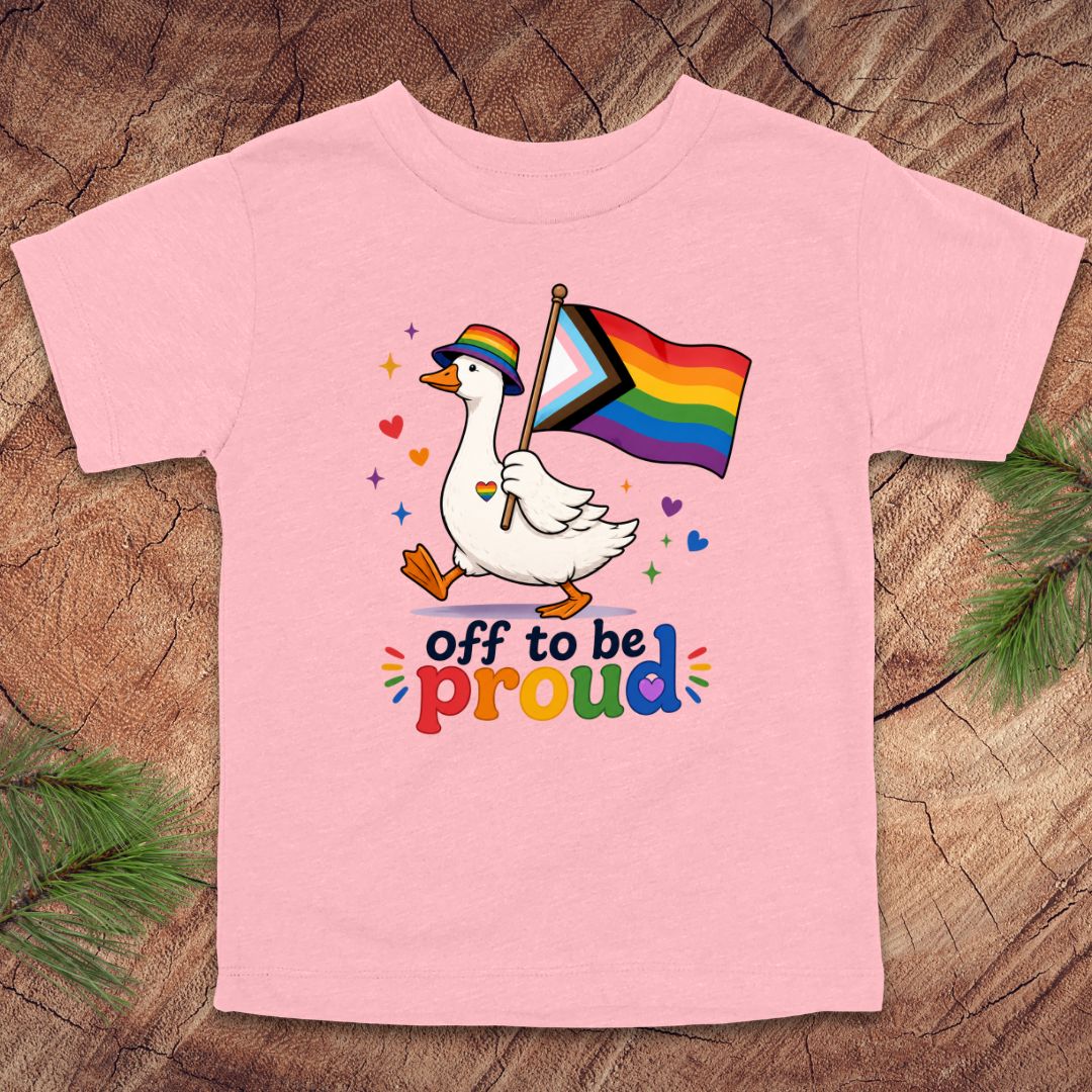 Pink t-shirt with a goose holding a rainbow flag and text 'off to be proud' on a wooden surface.