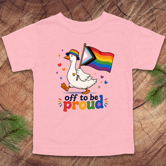 Pink t-shirt with a goose holding a rainbow flag and text 'off to be proud' on a wooden surface.