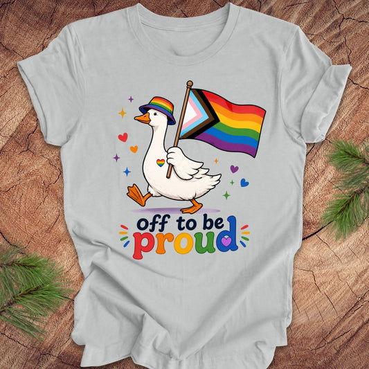 Gray t-shirt with a goose holding a rainbow flag and text 'off to be proud' on a wooden surface.
