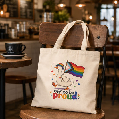Tote bag with a colorful design of a goose holding a rainbow flag, sitting on a chair in a café.