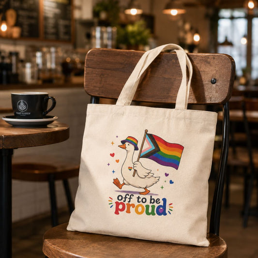 Tote bag with a colorful design of a goose holding a rainbow flag, sitting on a chair in a café.