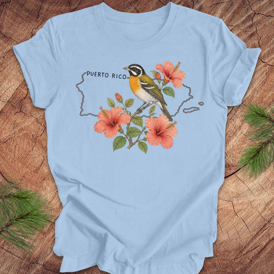 Light blue t-shirt with Puerto Rico outline and spindalis and hibiscus design on a wooden surface