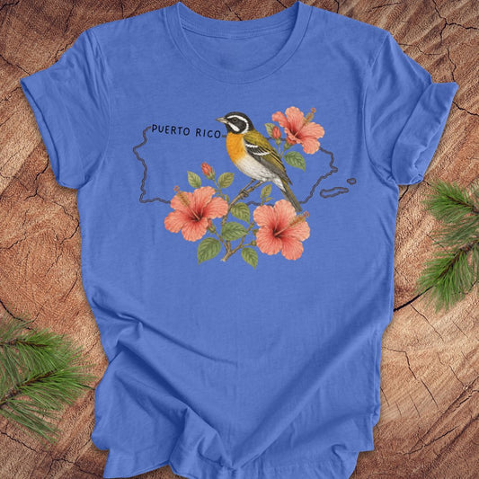 Blue t-shirt with Puerto Rico outline and spindalis and hibiscus design on a wooden surface