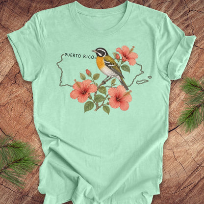 Mint green t-shirt with Puerto Rico outline and spindalis and hibiscus design on a wooden surface