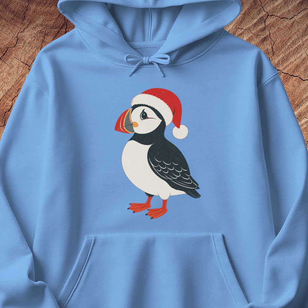 A light blue hoodie laid flat on a rustic wood background, featuring a printed illustration of a puffin wearing a red Santa hat on the chest.