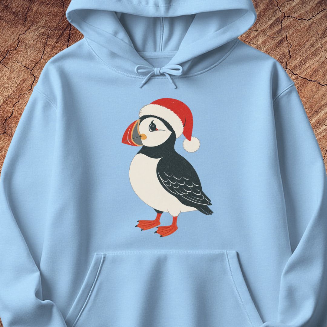 A light blue pullover hoodie featuring a graphic of a puffin wearing a red Santa hat, displayed flat against a textured brown wood background.