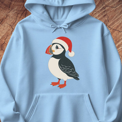 A light blue pullover hoodie featuring a graphic of a puffin wearing a red Santa hat, displayed flat against a textured brown wood background.