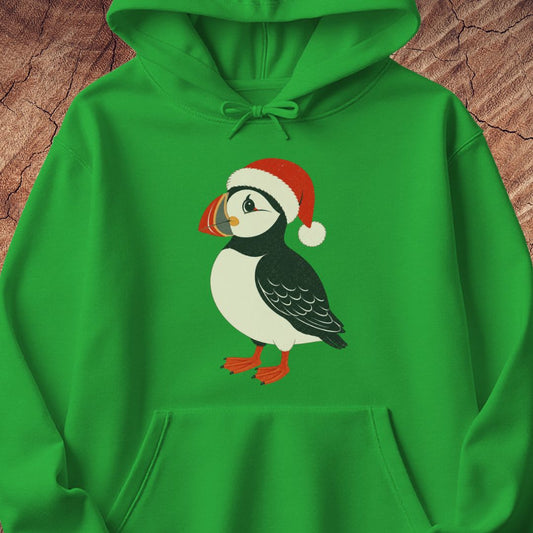 A bright green pullover hoodie featuring a cartoon puffin wearing a red Santa hat printed on the chest, laid flat on a textured brown wood background.