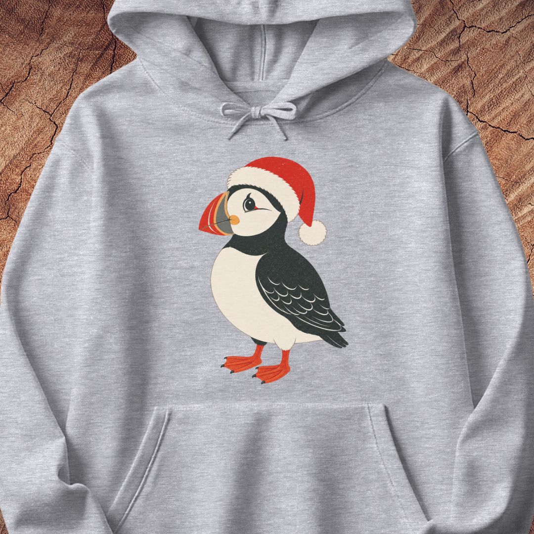 A light heather gray pullover hoodie featuring a cartoon puffin wearing a red Santa hat on the front, displayed against a rustic wood background.