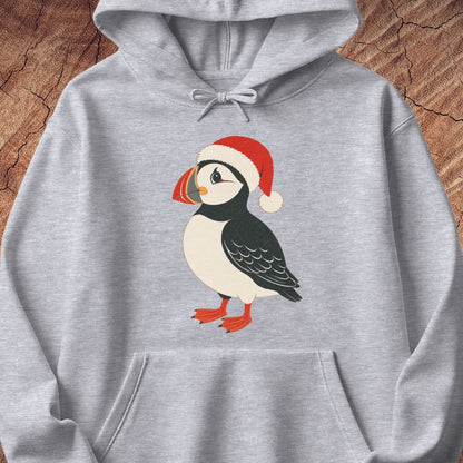 A light heather gray pullover hoodie featuring a cartoon puffin wearing a red Santa hat on the front, displayed against a rustic wood background.