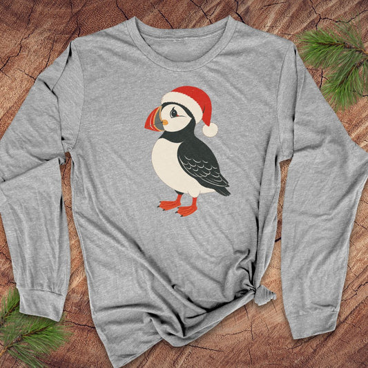 A heather gray long-sleeve t-shirt featuring a cartoon puffin wearing a red Santa hat, laid flat on a rustic wood background with pine needles in the corners.