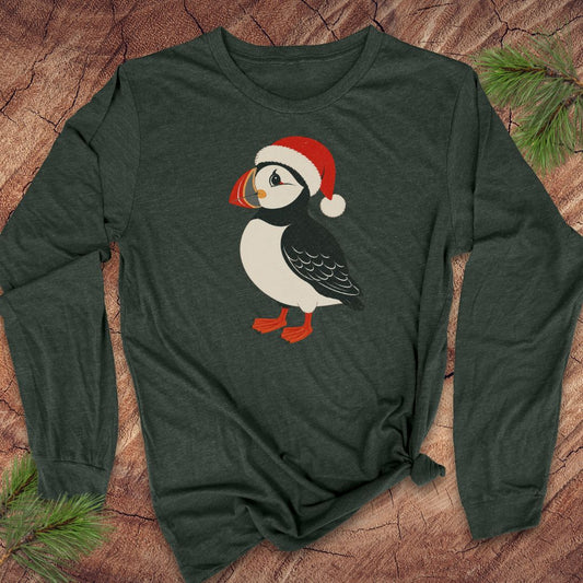 A dark forest green long-sleeve t-shirt with a graphic of a puffin wearing a red Santa hat, laid flat on a rustic wood background with pine branches.