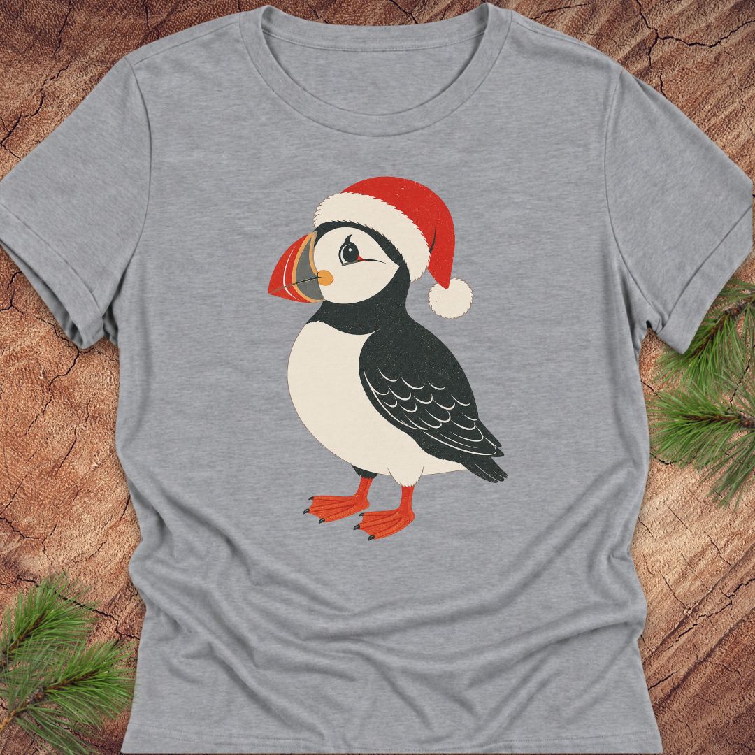 A heather gray short-sleeve t-shirt featuring a cartoon puffin wearing a red Santa hat, laid flat on a rustic wood background with pine branches in the corners.
