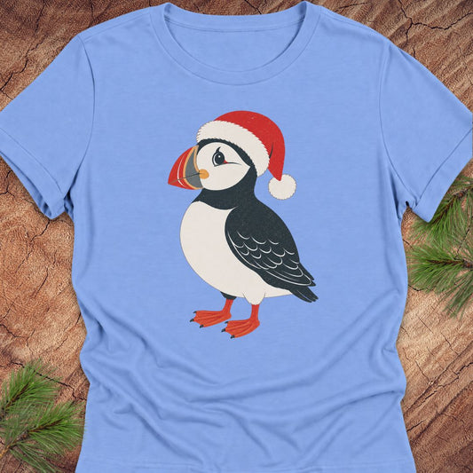 A light blue short-sleeve crewneck t-shirt featuring a cartoon puffin wearing a red Santa hat, laid flat on a rustic wood background with pine needles in the corners.