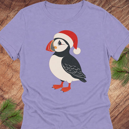 A lavender short-sleeve crewneck t-shirt featuring a cartoon puffin wearing a red Santa hat, laid flat on a rustic wood background with pine branches in the corners.