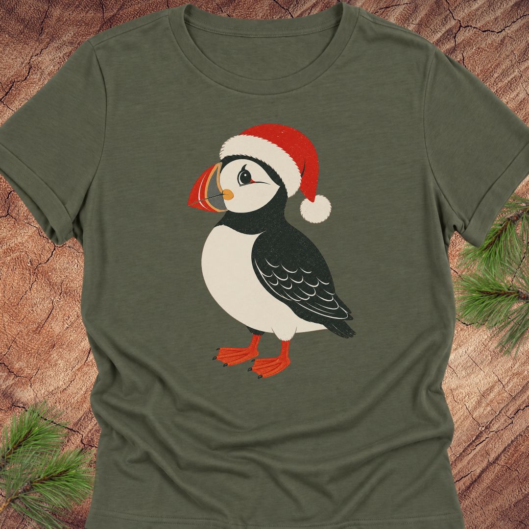 An olive green short-sleeve t-shirt featuring a cartoon puffin wearing a red Santa hat, laid flat on a rustic wood background with pine branches in the corners.