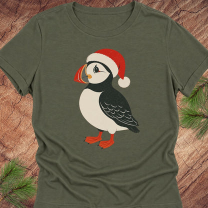 An olive green short-sleeve t-shirt featuring a cartoon puffin wearing a red Santa hat, laid flat on a rustic wood background with pine branches in the corners.