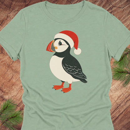 A sage green short-sleeve t-shirt featuring a cartoon puffin wearing a red Santa hat, laid flat on a rustic wood background with pine branches in the corners.