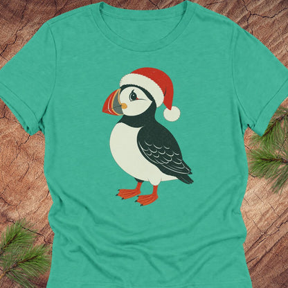 A teal short-sleeve crewneck t-shirt featuring a cartoon puffin wearing a red Santa hat, laid flat on a rustic wood background with pine branches in the corners.