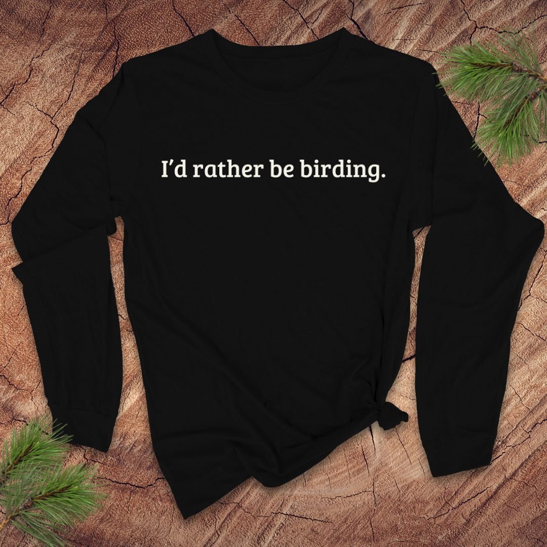 Black long-sleeve shirt with 'I'd rather be birding.' text on a wooden surface with pine branches.
