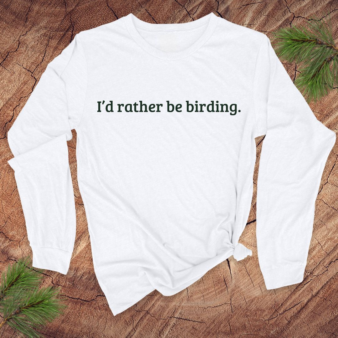 White long-sleeve shirt with 'I'd rather be birding.' text on a wooden surface with pine branches.