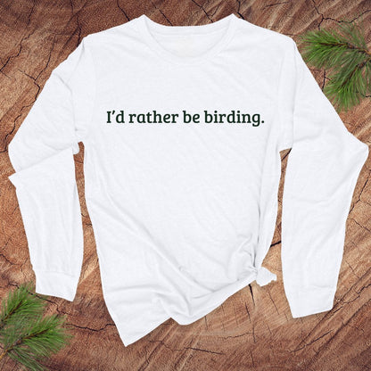 White long-sleeve shirt with 'I'd rather be birding.' text on a wooden surface with pine branches.