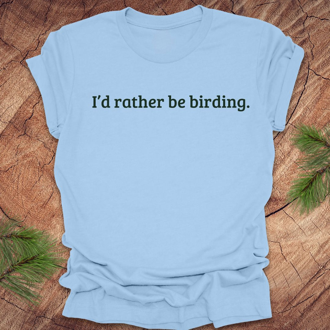 Light blue t-shirt with 'I'd rather be birding.' text on a wooden surface with greenery.
