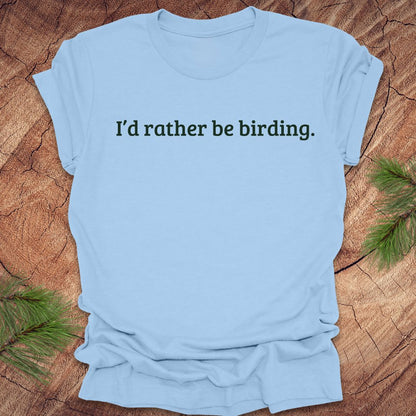 Light blue t-shirt with 'I'd rather be birding.' text on a wooden surface with greenery.