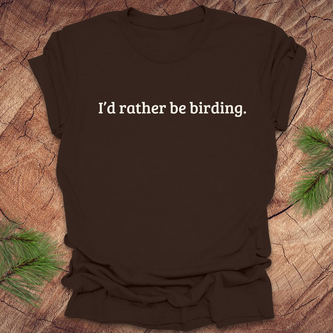 Brown t-shirt with 'I'd rather be birding.' text on a wooden surface with pine branches.