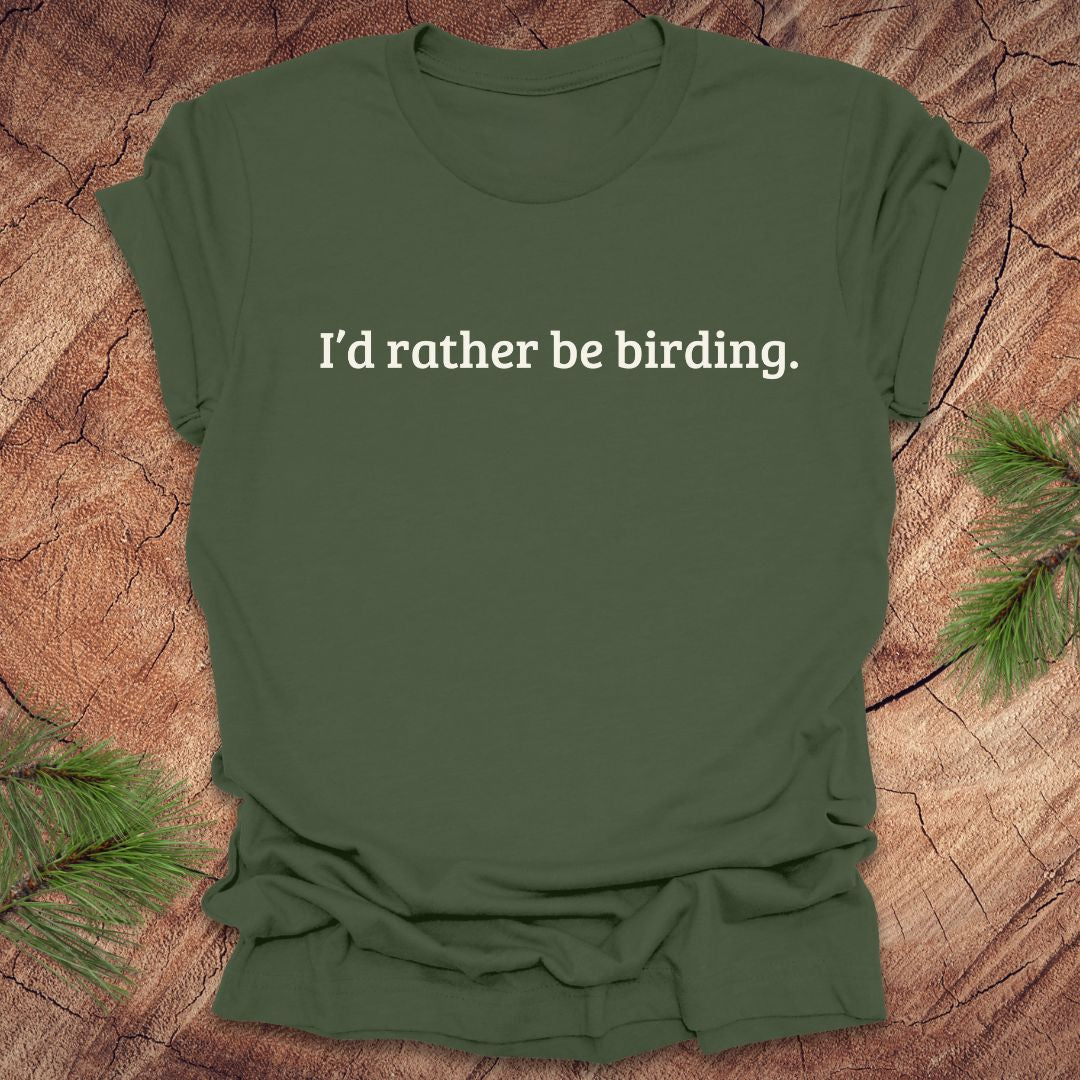 Green t-shirt with 'I'd rather be birding.' text on a wooden surface with pine branches.