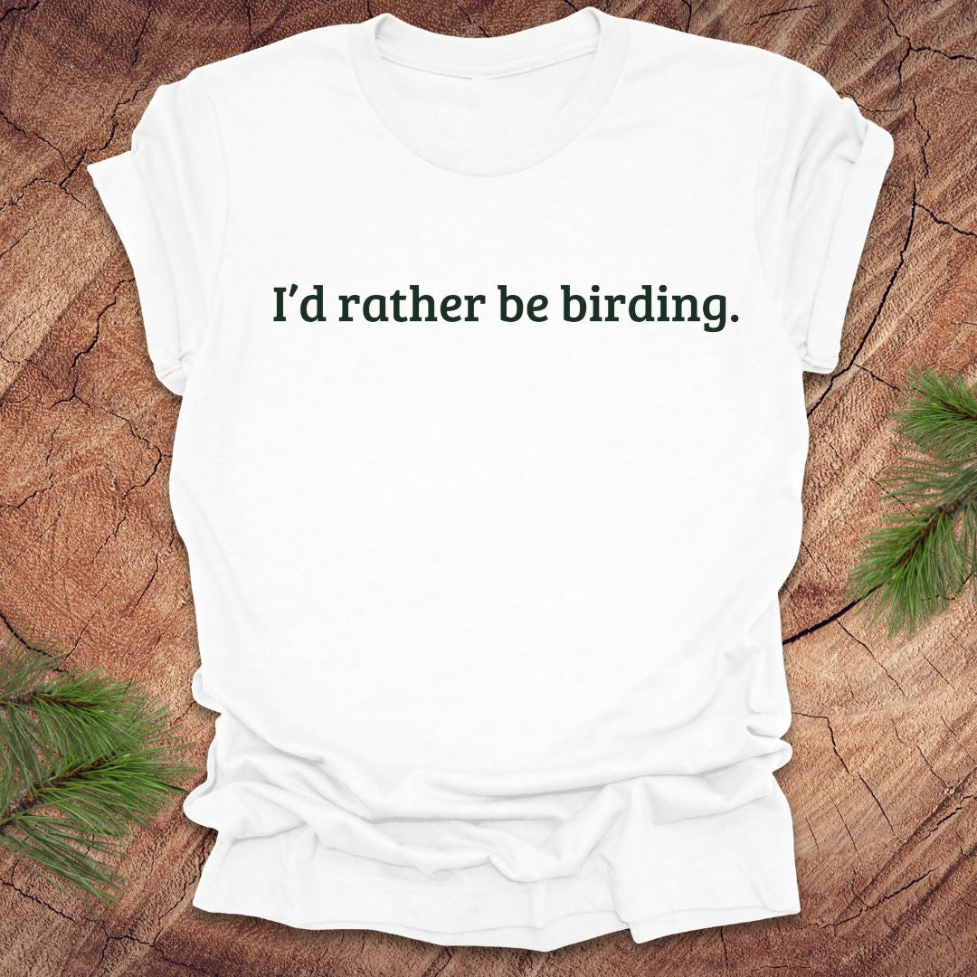 White t-shirt with 'I'd rather be birding.' text on a wooden surface with greenery.