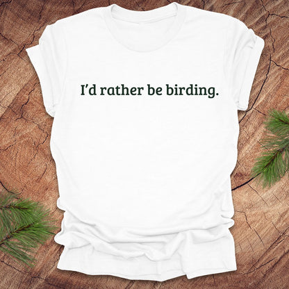 White t-shirt with 'I'd rather be birding.' text on a wooden surface with greenery.