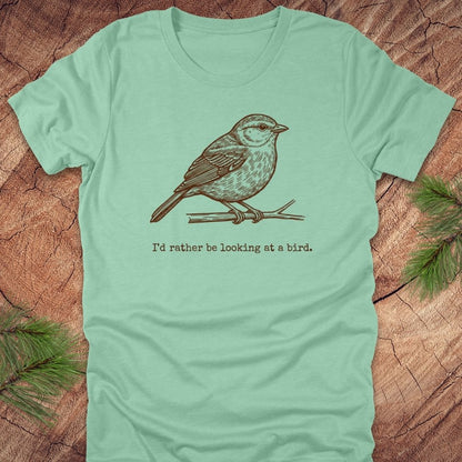 Mint green t-shirt with a hand drawn brown bird on a branch with text, 'i'd rather be looking at a bird.' on a wooden background.