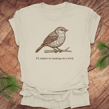 Beige t-shirt with a hand drawn brown bird on a branch with text, 'i'd rather be looking at a bird.' on a wooden background.