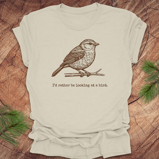 Beige t-shirt with a hand drawn brown bird on a branch with text, 'i'd rather be looking at a bird.' on a wooden background.