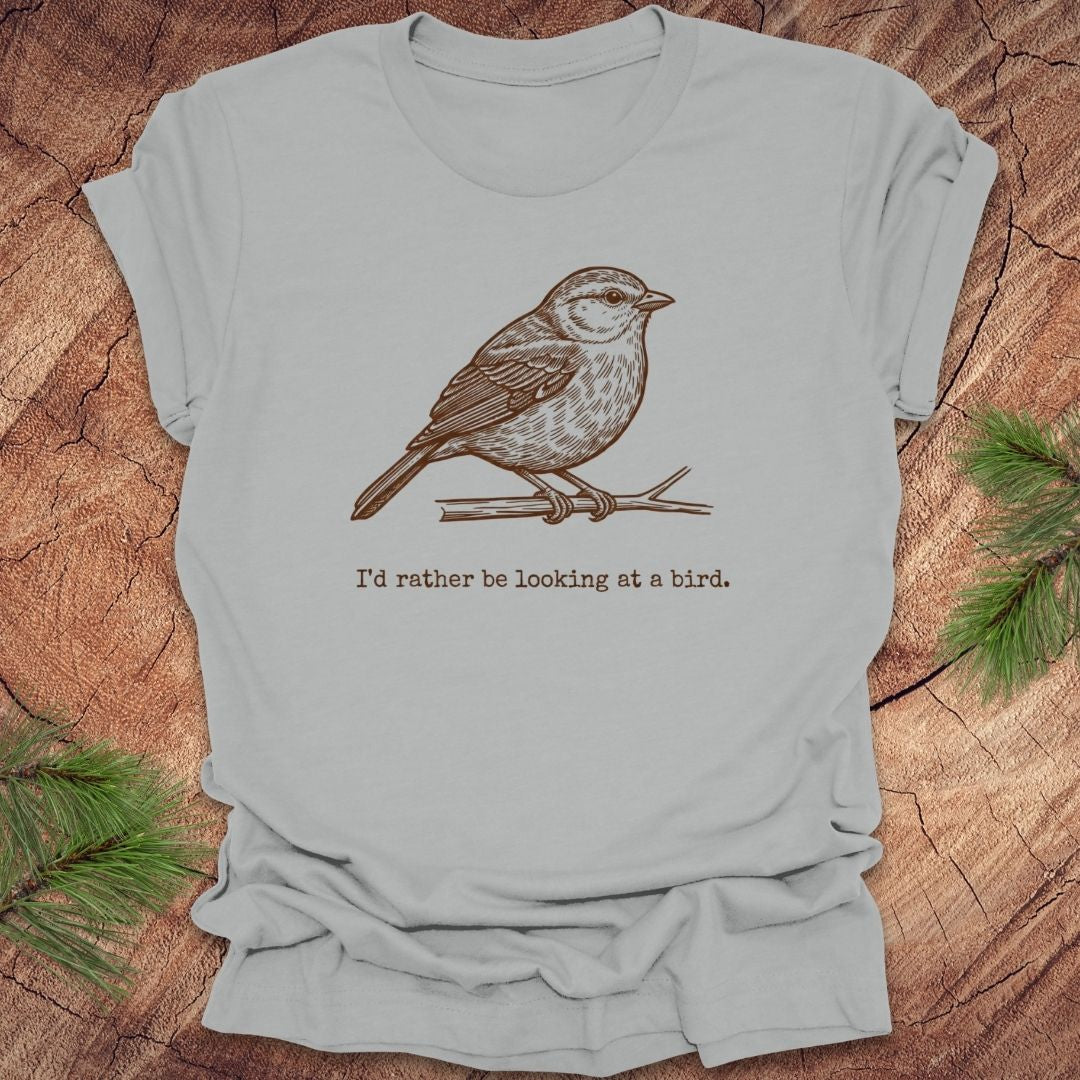 Silver t-shirt with a hand drawn brown bird on a branch with text, 'i'd rather be looking at a bird.' on a wooden background.