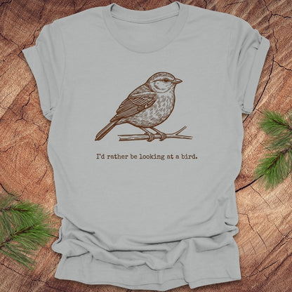 Silver t-shirt with a hand drawn brown bird on a branch with text, 'i'd rather be looking at a bird.' on a wooden background.