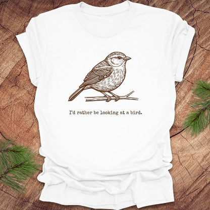 White t-shirt with a hand drawn brown bird on a branch with text, 'i'd rather be looking at a bird.' on a wooden background.