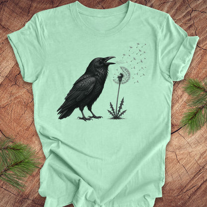 Mint green t-shirt with a raven and dandelion design on a wood background.