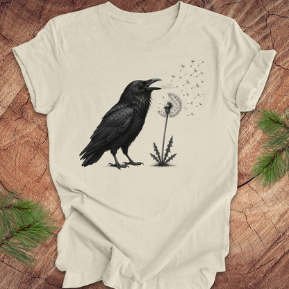 Beige t-shirt with a raven and dandelion design on a wood background.