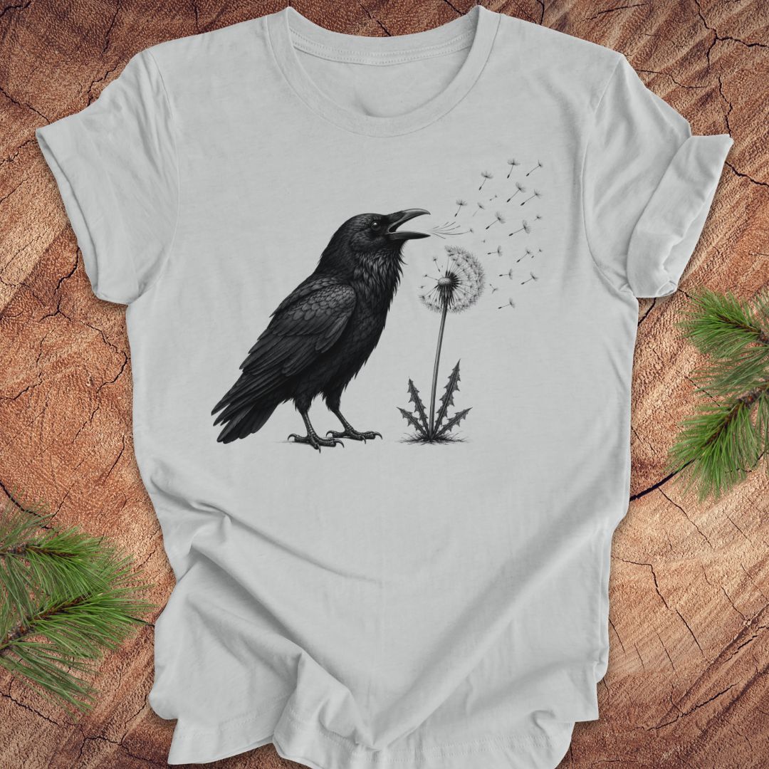 Gray t-shirt with a raven and dandelion design on a wood background.