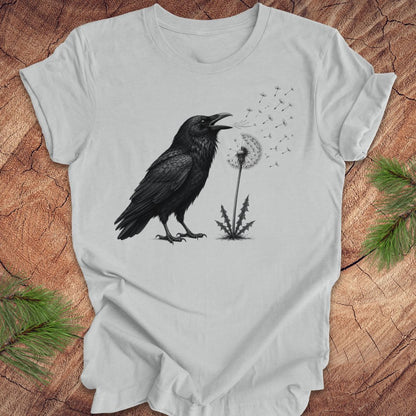 Gray t-shirt with a raven and dandelion design on a wood background.