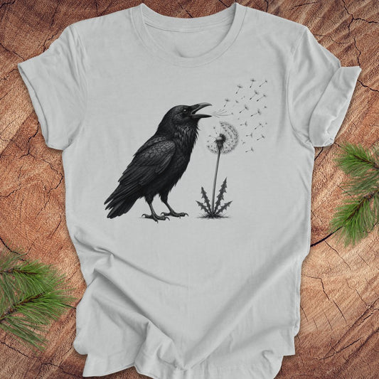 Gray t-shirt with a raven and dandelion design on a wood background.