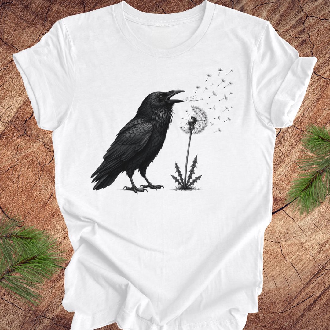 White t-shirt with a raven and dandelion design on a wood background.
